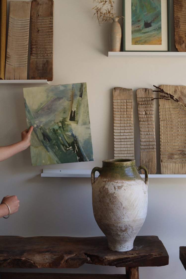 Person adjusting a painting on a wall with a rustic vase and wooden panels in the foreground.