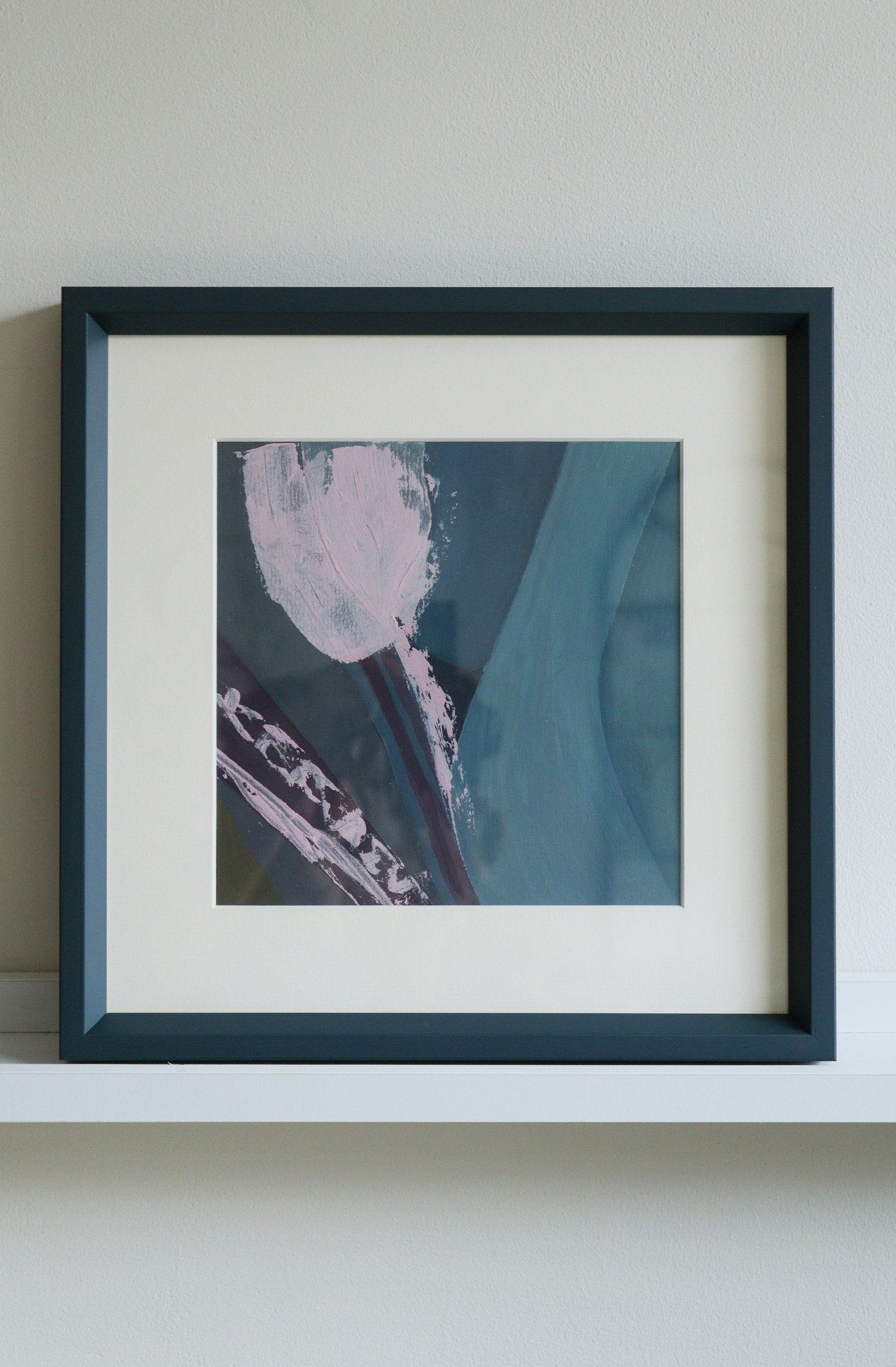 Framed abstract artwork with a blue and pink colour scheme on a white shelf.
