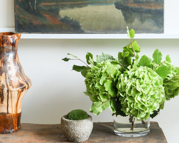 Decorative setup with a painting, vase, and green flowers on a wooden surface.