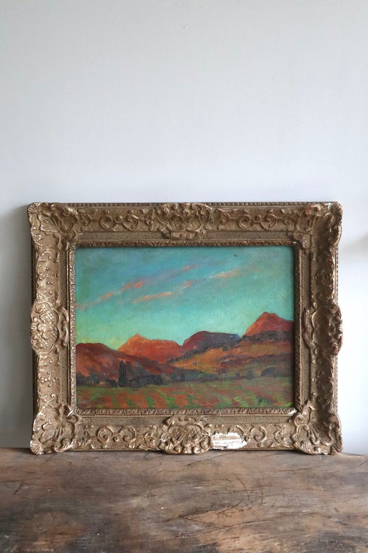 Vintage landscape painting in an ornate gold frame on a wooden surface.