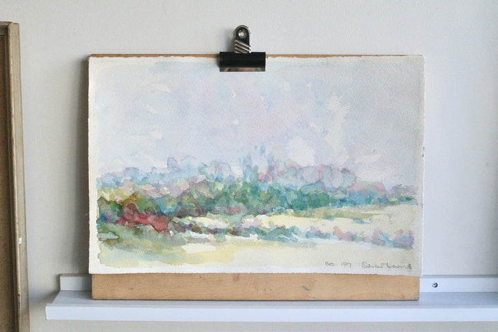 Watercolor landscape painting on a wooden board with a clip, against a white wall.