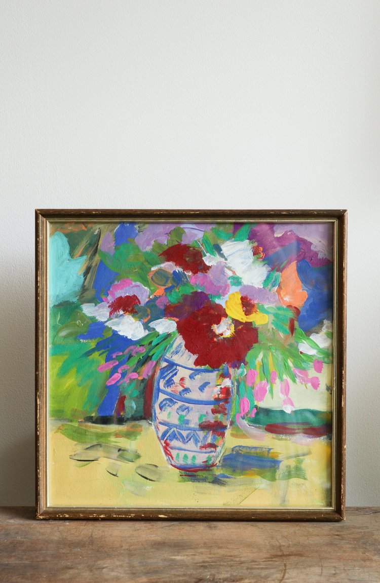 Framed painting of a colorful floral arrangement on a wooden surface with a white wall background.