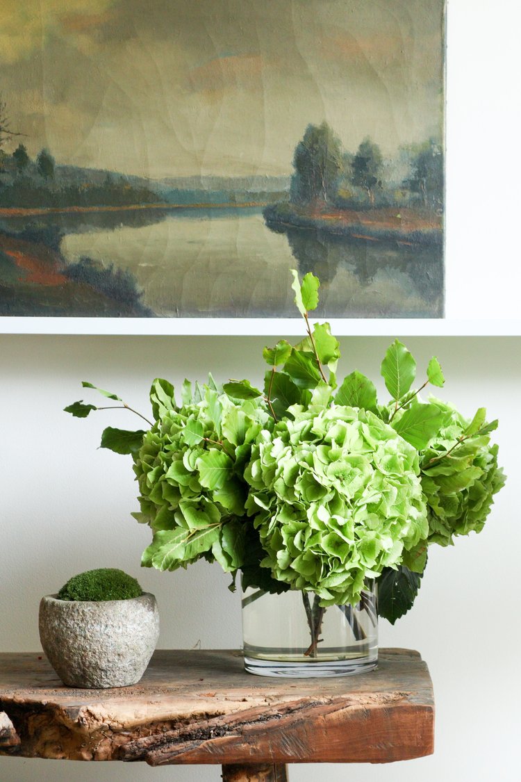Green flowers in a vase on a wooden table with a painting of a landscape in the background.