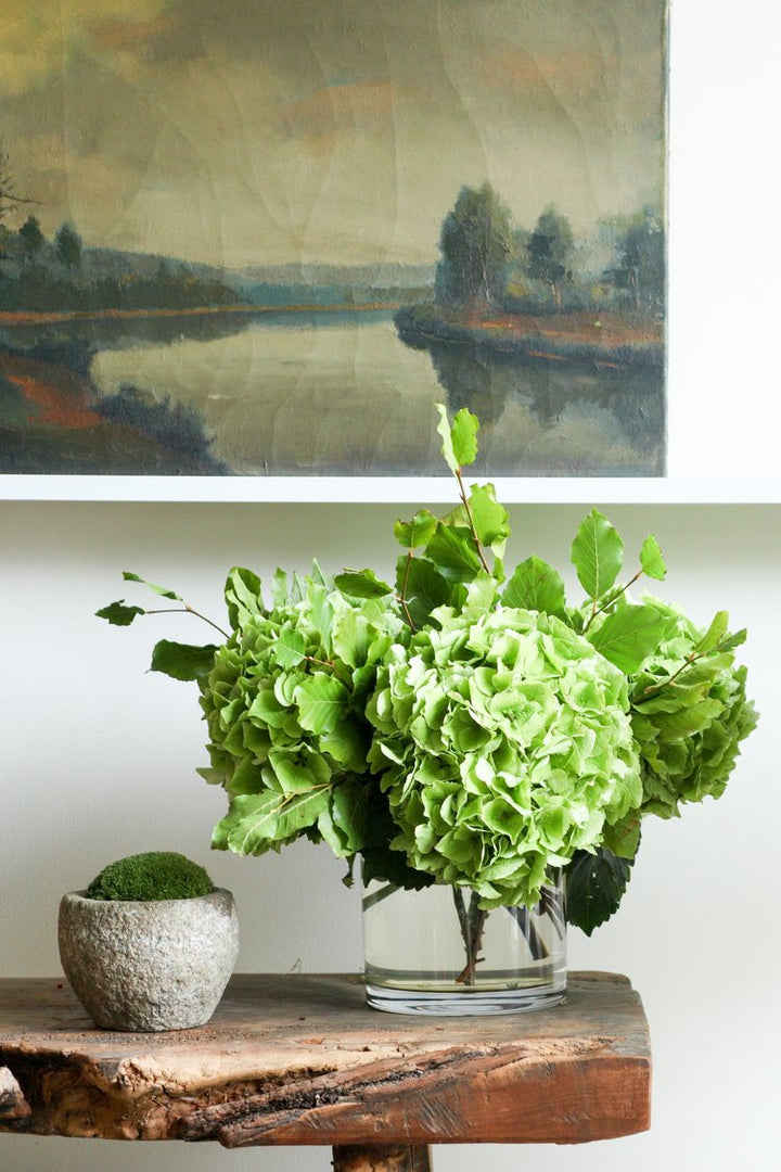 Green flowers in a vase on a wooden table with a painting of a landscape in the background.