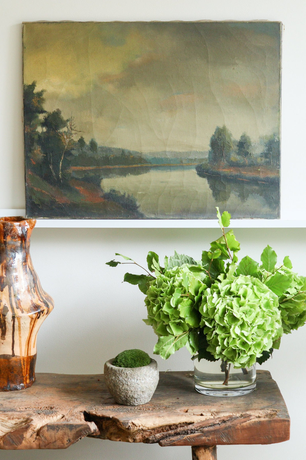 Decorative setting with a painting of a landscape, vase, and green flowers on a wooden surface.
