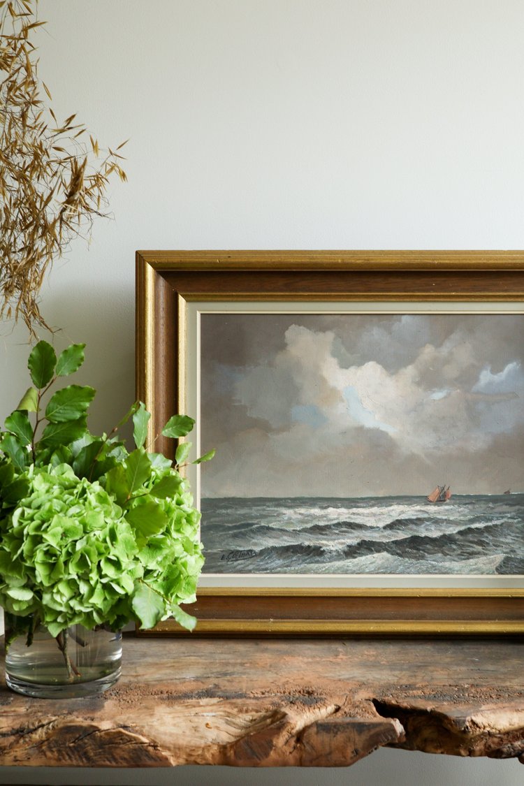 Framed painting of a seascape with a boat on a wooden shelf next to a green plant.