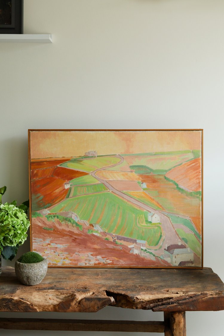 Framed landscape painting on a wooden table with a plant in the corner.