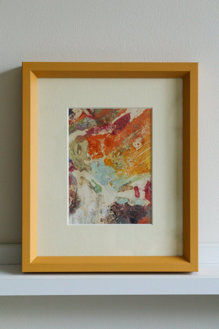 Framed abstract artwork with a colorful painting inside on a white surface.