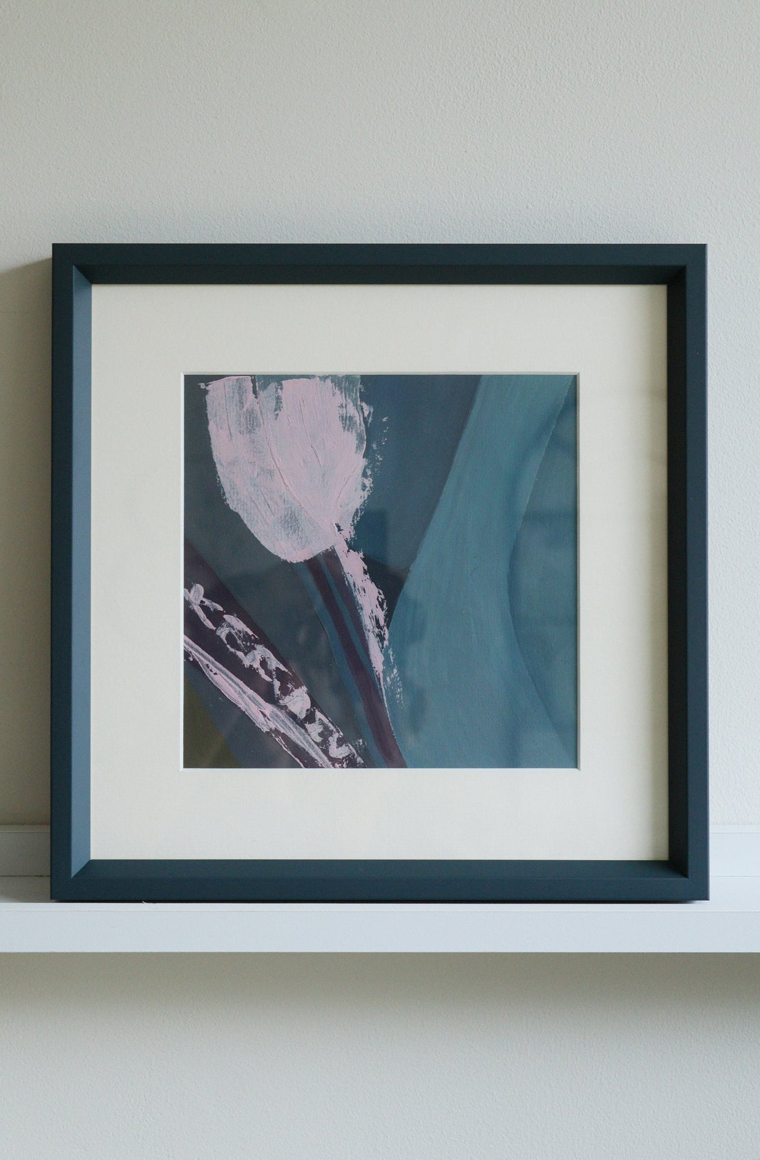 Framed abstract artwork with a blue and pink color scheme on a white shelf.