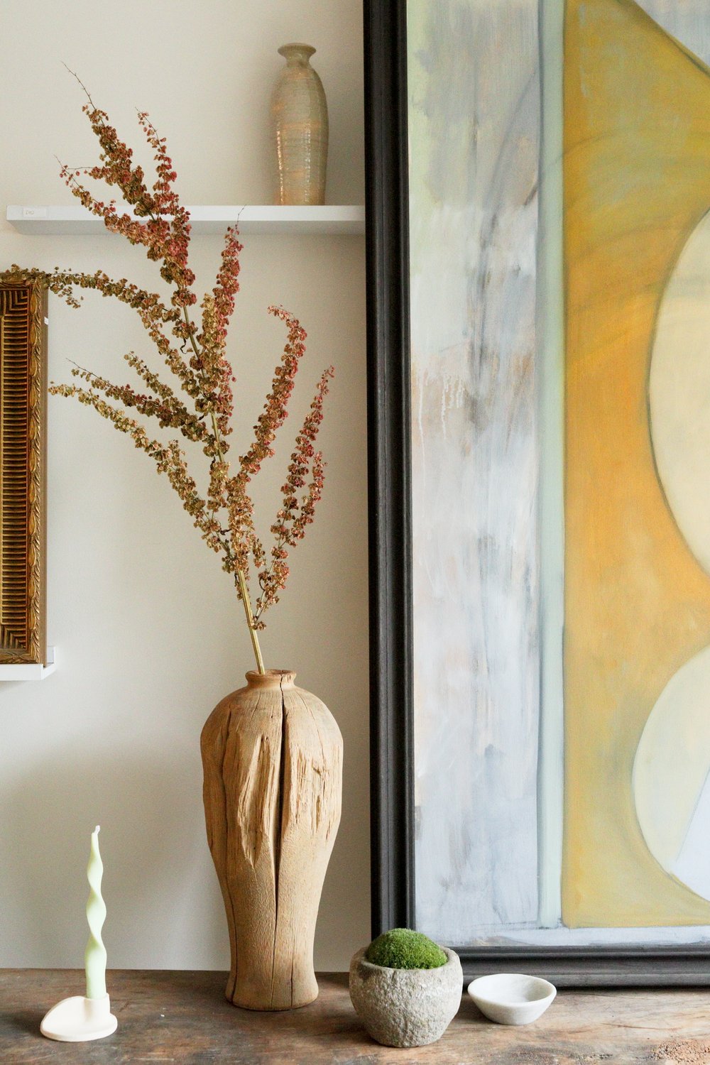 Decorative setting with a wooden vase, dried plants, and a painting in a room.