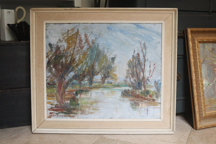 Framed painting of a landscape with trees and water on a wooden floor.