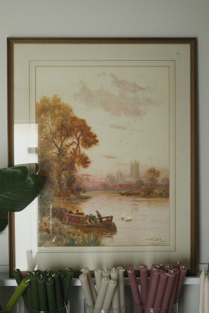 Framed landscape painting of a river scene with trees and buildings on a wall.