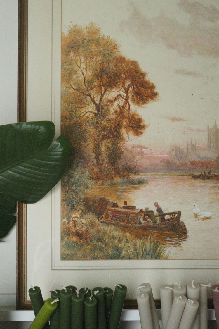 Framed painting of a river scene with a boat and trees, placed on a shelf with plants and candles.