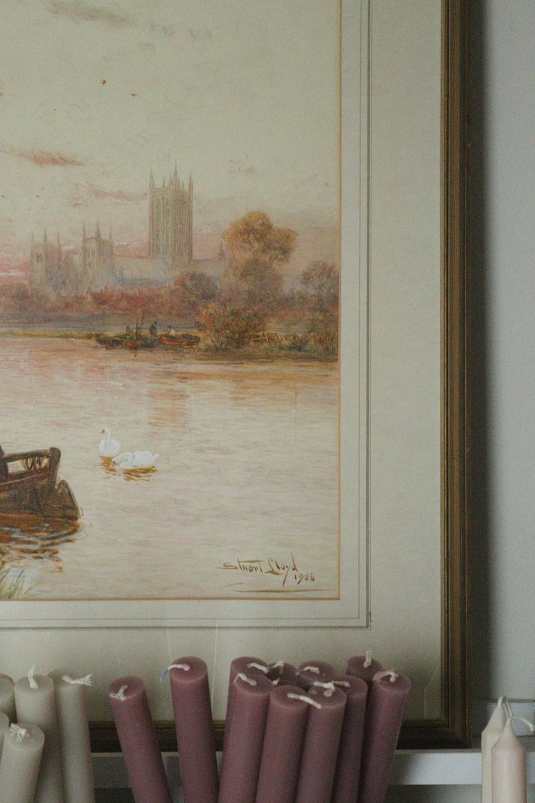 Framed painting of a landscape with a river and buildings, below which are candles.
