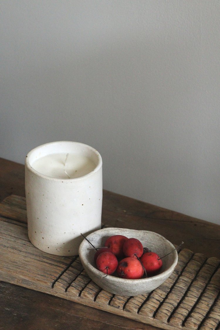 White candle in a textured container on a wooden surface with a small bowl of red fruits.