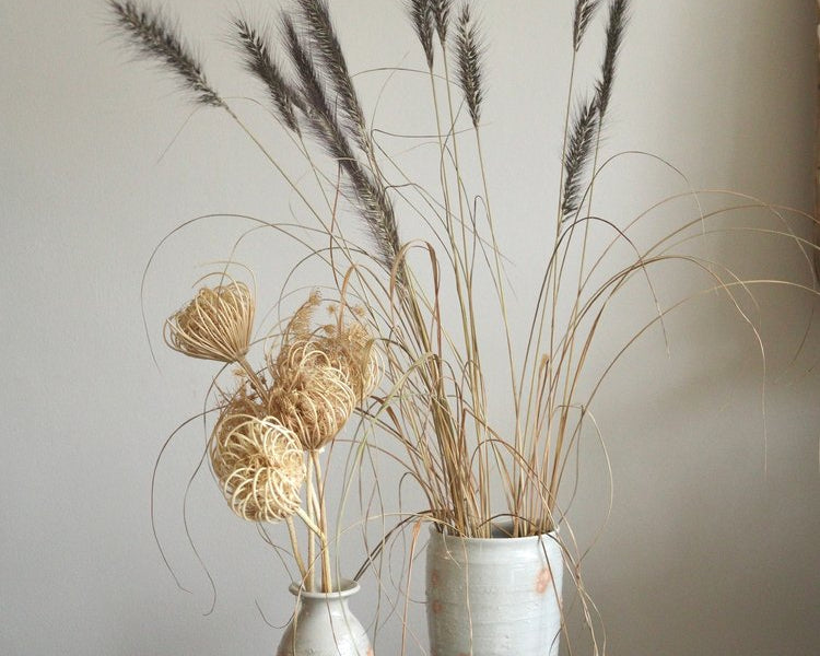Two ceramic vases with dried plants on a wooden surface against a plain wall.