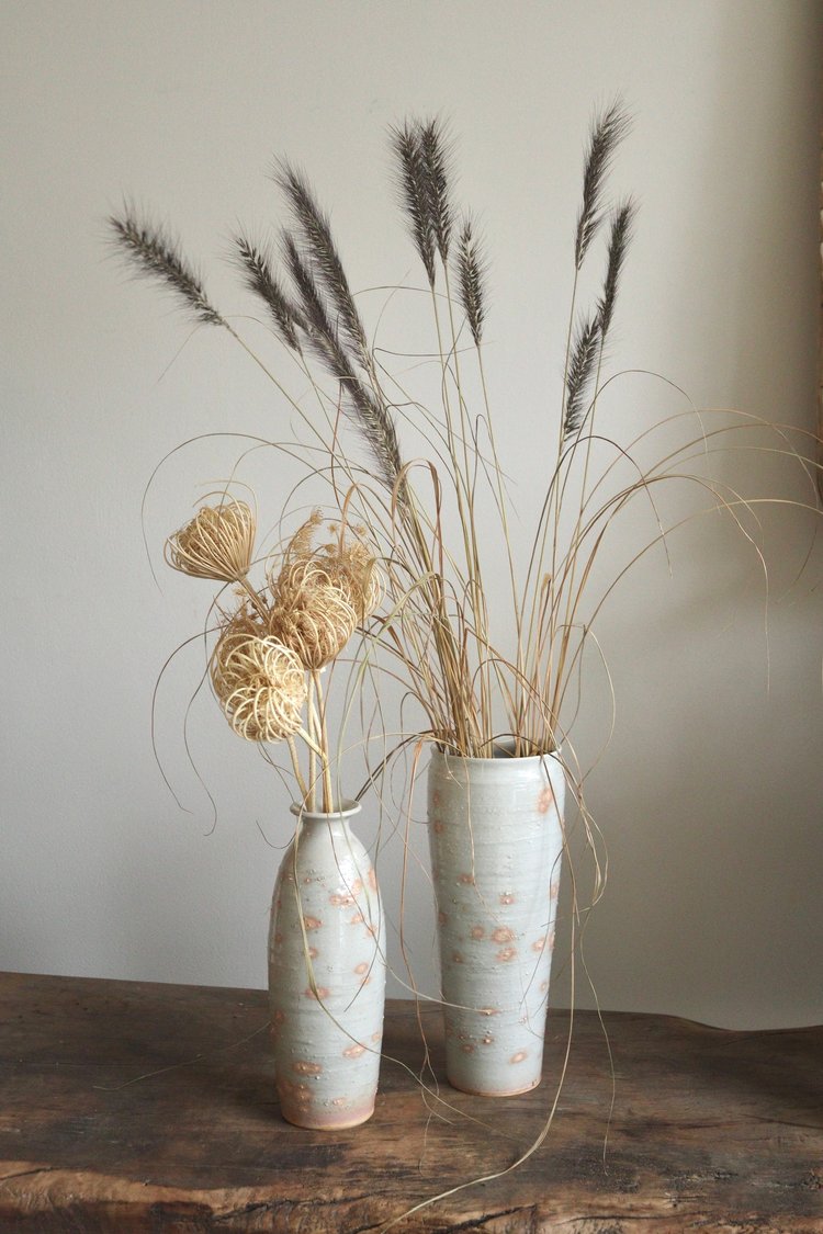 Two ceramic vases with dried plants on a wooden surface against a plain wall.