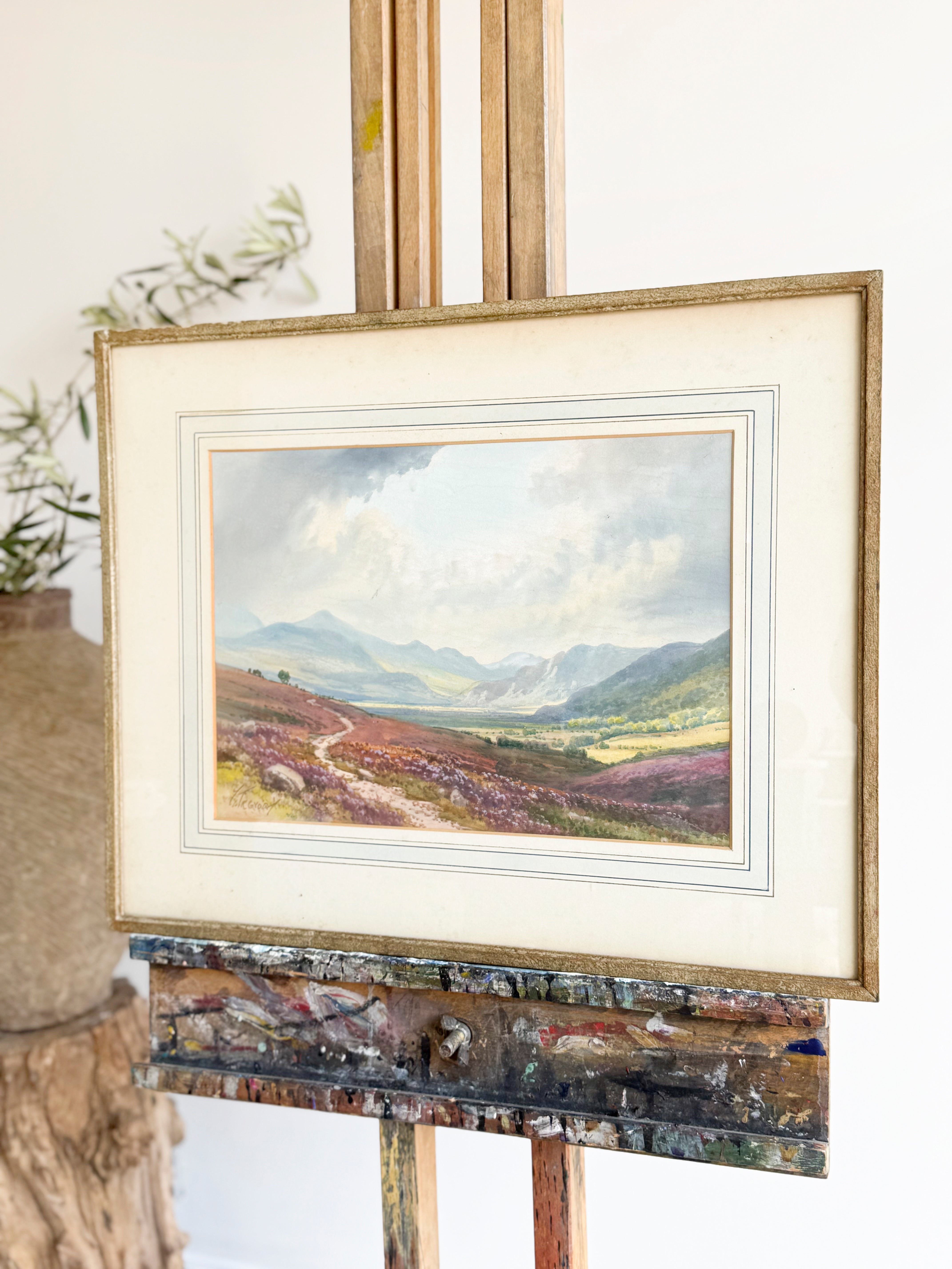 Framed landscape painting on an easel with a scenic view of mountains and fields.