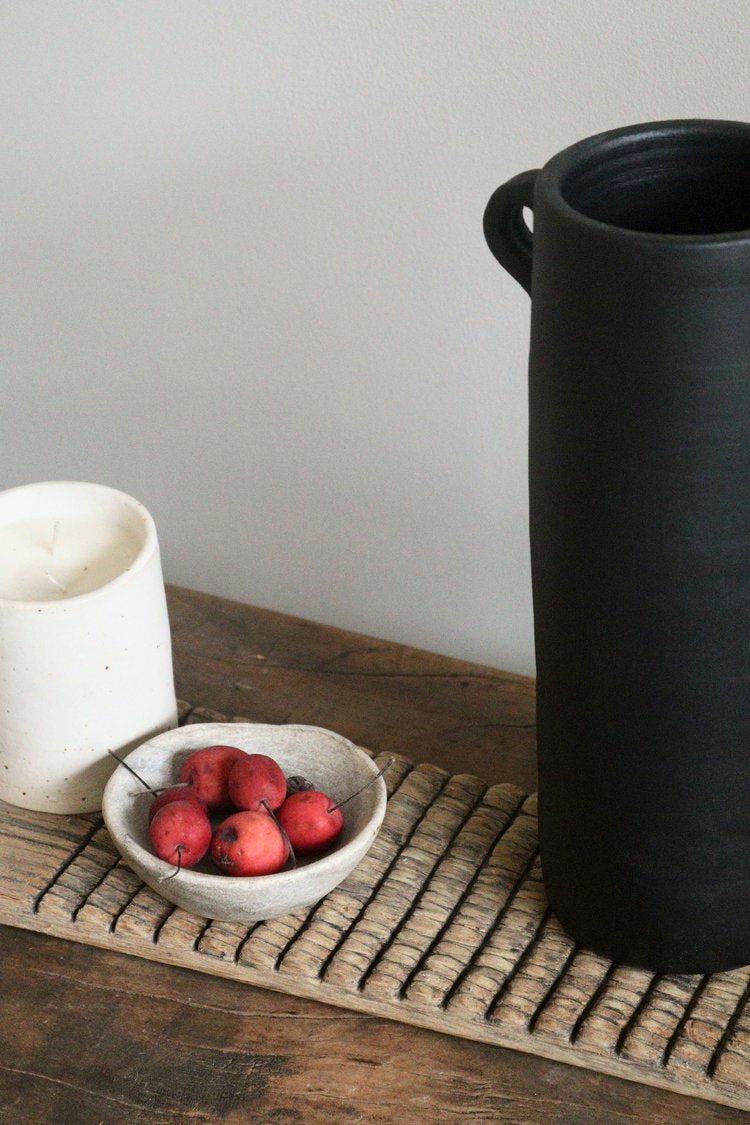 Black vase on a wooden surface with a bowl of red apples and a white container.
