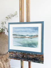 Framed artwork of a coastal scene on an easel with a white background