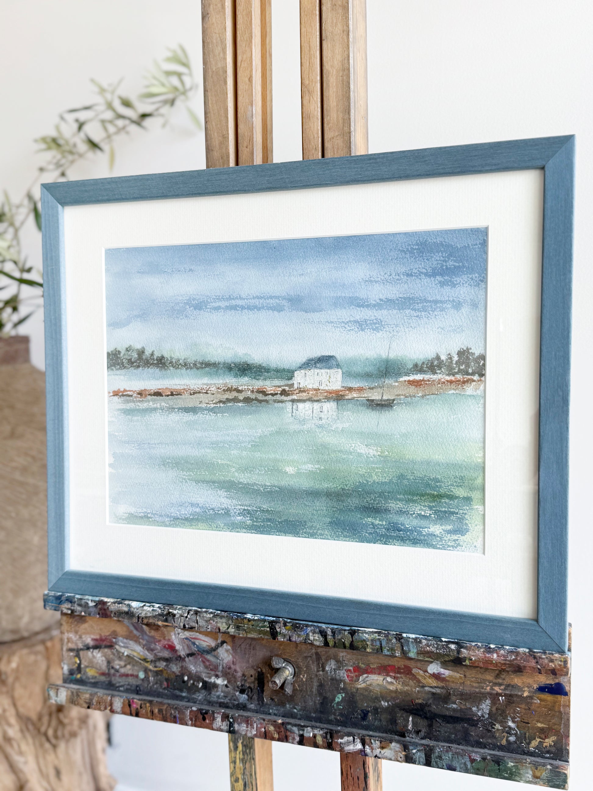 Framed watercolor painting of a coastal scene on a wooden easel.